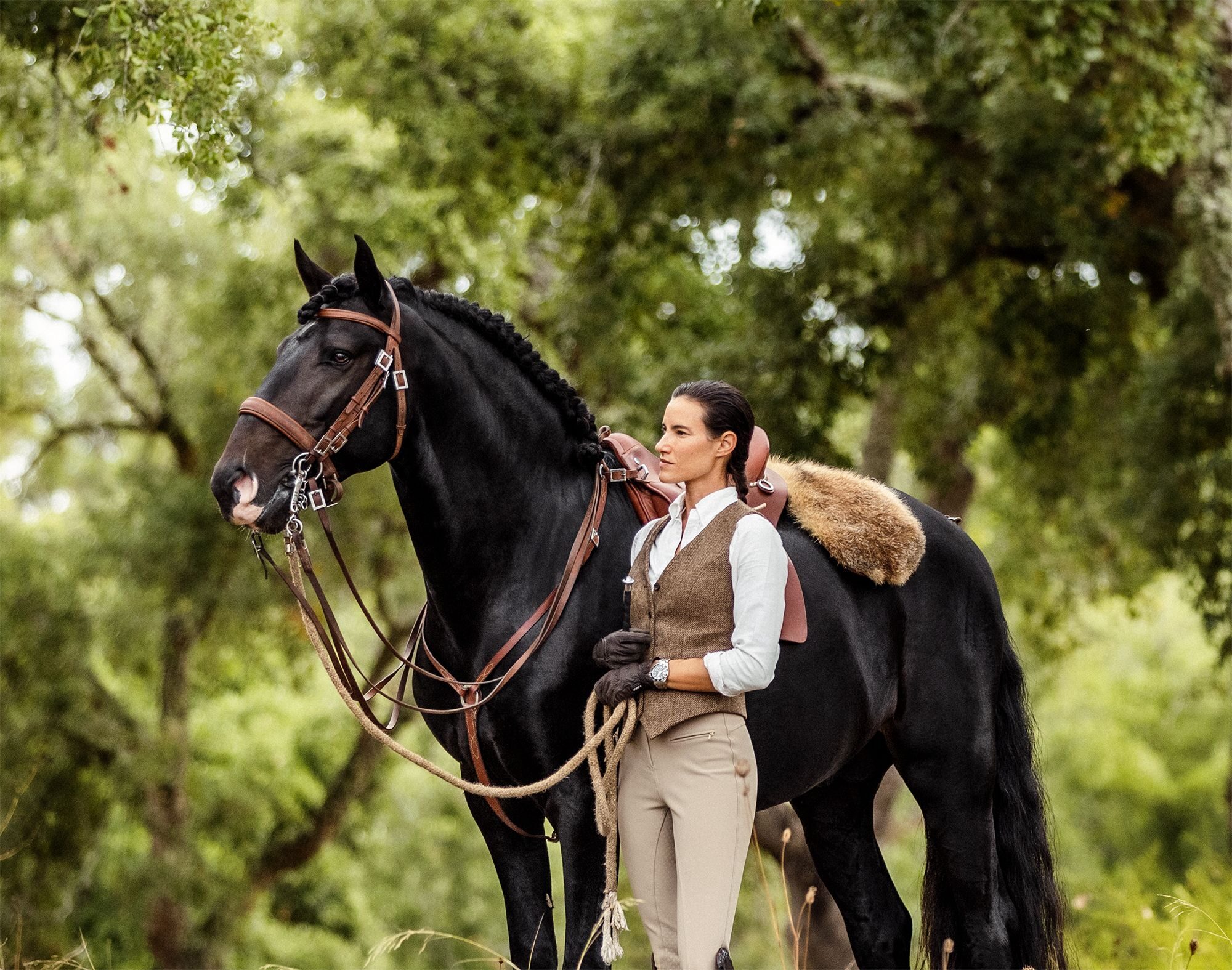 A rider in equestrian attire stands quietly beside her black Lusitano in the cork forest at AMAIA Valley.